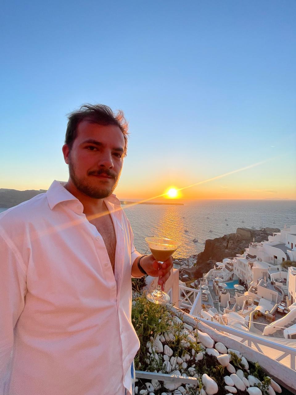Gabriel ao pôr do sol em Oia, Santorini, com drink na mão